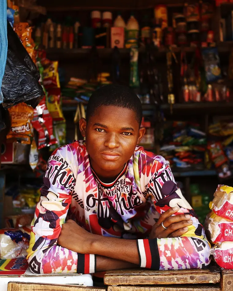 Un jeune commerçant accoudé au comptoir de son kiosque rempli de produits.