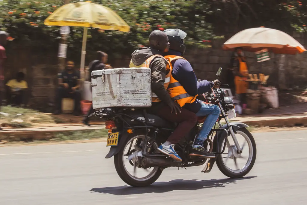 Un livreur à moto transporte une caisse de marchandises dans une rue animée.