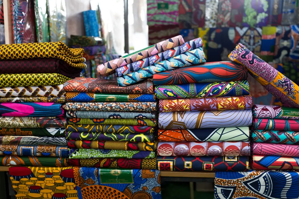 Des piles de tissus wax aux motifs colorés, sur un étal de marché.